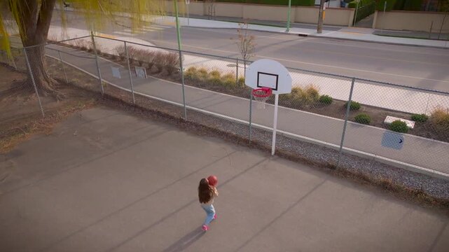 Aerial shot of a young girl with long brown hair standing in front of a basketball hoop at a public court along a street, aiming to shoot, but missed twice and made the shot on the third attempt. 