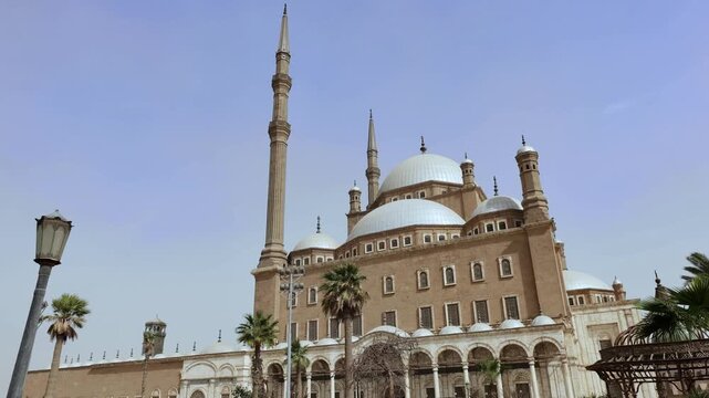 Stunning Ottoman mosque with gleaming white domes and towering minaret at Cairo Citadel, Egypt, elegant arched gallery and palm trees in sunny clear weather.