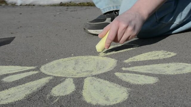 Woman drawing yellow flower on asphalt with chalk. Girl kneeling on ground creating creative street art. Childhood pastime outdoors during summer sunny day time.