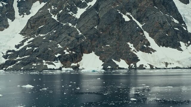 Slow sideways pan along dramatic jagged snow-patched Antarctic rocky cliff coastline with dark reflective icy ocean and brash ice