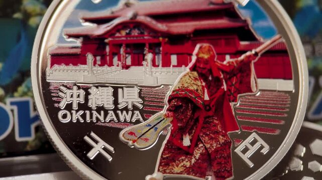 Close up video of an Okinawa prefectural 1000 yen silver proof coin featuring a polished surface, colorful regional design, traditional figure, floral elements, and geometric details.