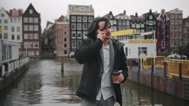 Young businessman talking on the phone and holding coffee while walking on a bridge in amsterdam on a windy day
