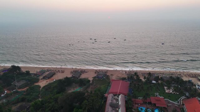 Drone shot capturing a calm seaside sunset with warm pastel tones spreading across the sky, reflecting on rippling waters while the shoreline and coastal homes sit in peaceful silhouette.