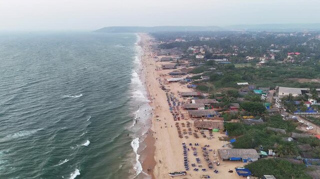 Cinematic aerial view of Baga Beach stretching along the coastline, with waves rolling onto golden sands and rows of beach shacks and sunbeds lining the shore, capturing a lively and vibrant seaside
