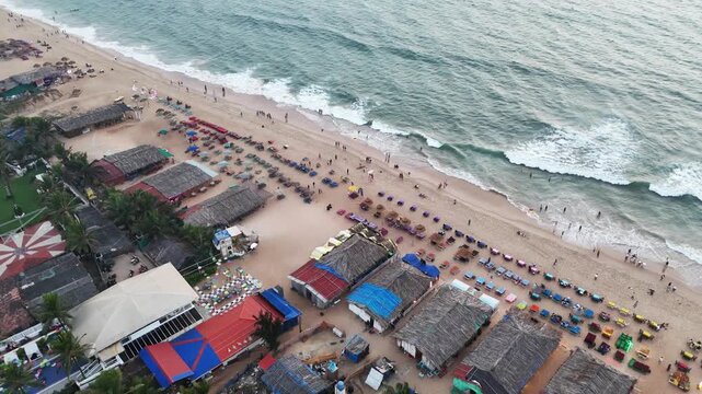 Cinematic aerial view of a vibrant beach with neatly arranged sunbeds and colorful umbrellas, as waves crash rhythmically along the shoreline. People stroll and relax, creating a lively coastal scene.