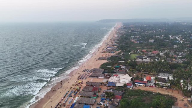 Drone shot capturing a dynamic coastline from above, where textured ocean waves meet a busy beach lined with shacks and seating rows. The contrast between movement and structure creates a cinematic.