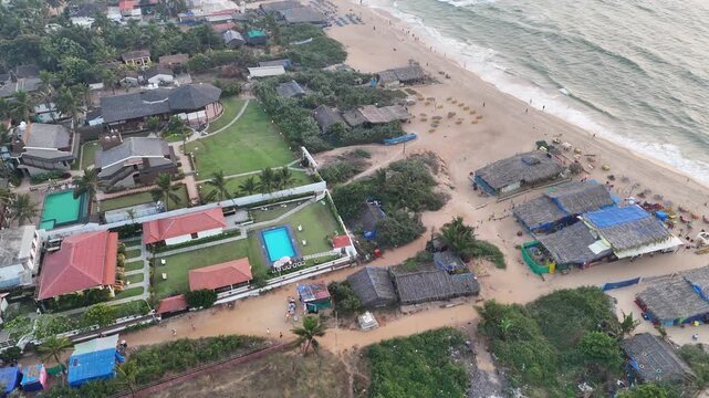 Cinematic aerial view of a beachfront resort near Baga Beach, featuring landscaped lawns, a bright blue pool, and tiled villas, seamlessly blending with nearby beach shacks and the sandy shoreline.