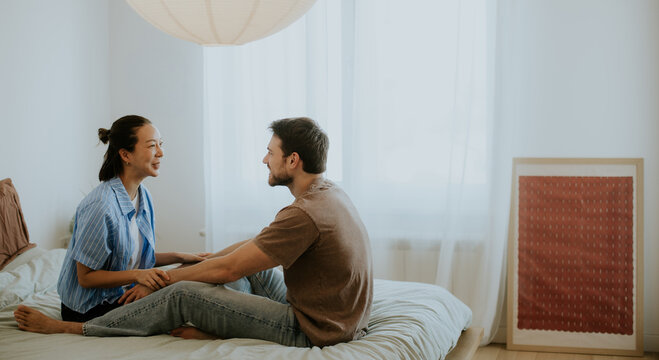 Korean woman and Caucasian man sitting and talking in a bedroom with natural light