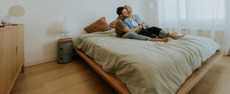 A Korean woman and a Caucasian man sit side by side on a bed in a bright bedroom