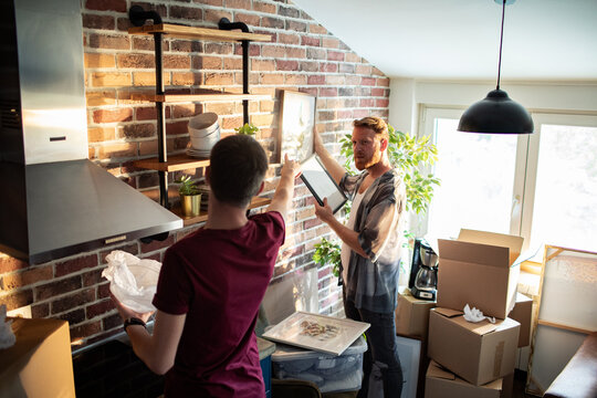 Two men hang artwork while unpacking in a brick kitchen
