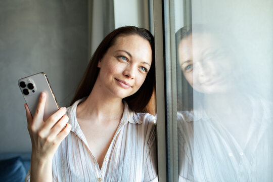 Smiling woman holding smartphone by window at home