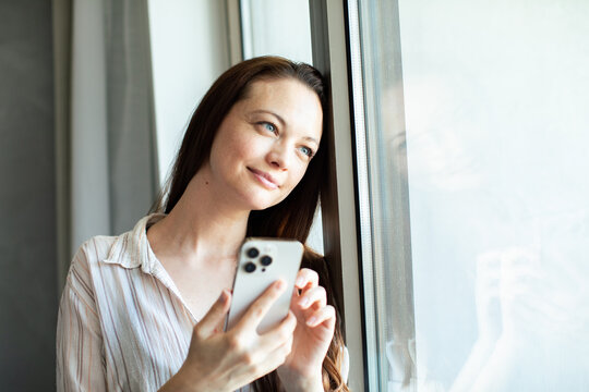 Young woman holding smartphone by window at home
