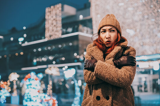 Woman feeling cold and uncomfortable while standing outdoors in a city at night, surrounded by festive lights