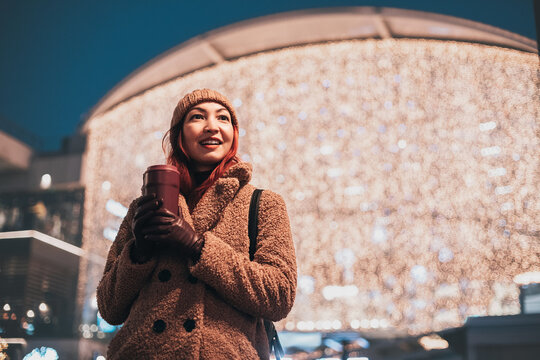 Young woman smiling, holding a coffee cup, surrounded by blurred city lights at night, embodying urban winter lifestyle