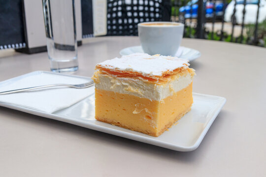 The original Bled cream cake (Cremeschnitte) on white plate and cup of cappuccino on a table. Traditional Slovenian dessert