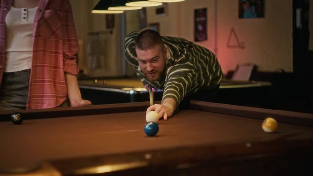 Tracking shot of young adult man hitting ball on billiards table while playing pool together with woman and discussing game at snooker hall