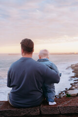 Fototapeta premium Father holding baby and looking at the sea. Calm emotional moment of parenting, family bonding, and peaceful lifestyle by the water.