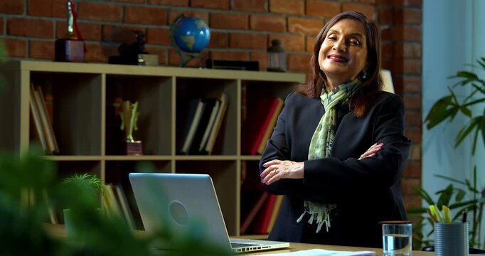 Senior Indian businesswoman in corporate attire working on laptop in cozy office space looks at camera with a smile then poses with folded hands and a thumbs up experienced female executive