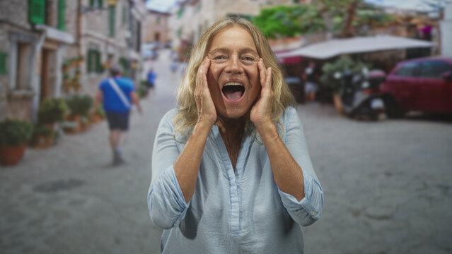Senior woman with blonde hair clasping hands to her chest and smiling with eyes closed on a cobbled street with cafe tables and potted plants; joy.