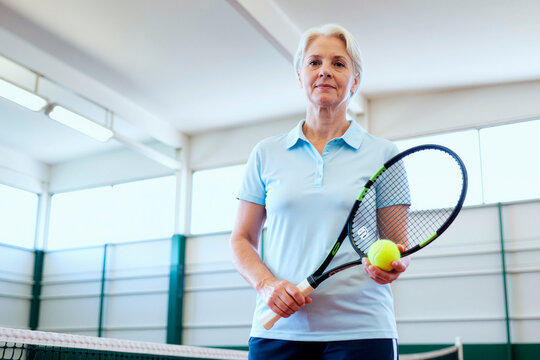 Portrait of senior Caucasian woman holding tennis racket and ball, standing on indoor court, promoting active aging and healthy lifestyle. Useful for fitness, wellness, senior sports marketing
