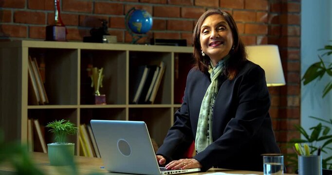 Senior Indian businesswoman in corporate attire working on laptop in cozy office space looks at camera with a smile then poses with folded hands and a thumbs up experienced female executive
