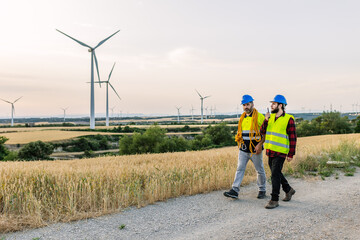 Obraz premium Engineers in hard hats and safety vests walking on a dirt road, discussing operations at a wind farm generating clean energy. Renewable energy and industrial workforce concept