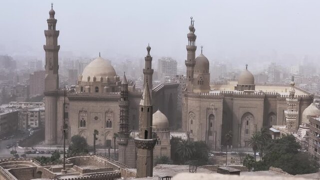 Dusty atmospheric skyline of Cairo, Egypt, dominated by the medieval Sultan Hassan and Al-Rifa'i mosque complex, featuring tall minarets, stone domes and ornate facades.
