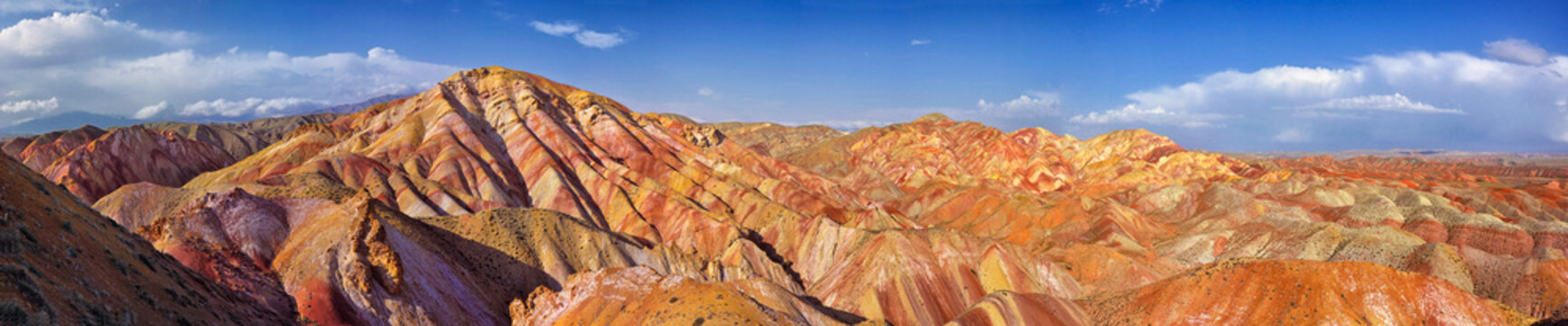 Panoramic view of colorful striped mountains with diagonal layers in red yellow orange green purple tones, dramatic light and shadow under blue sky