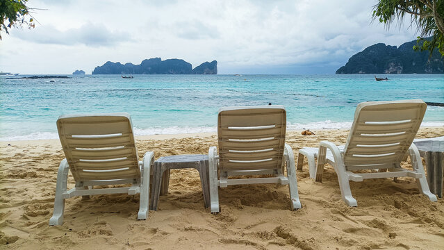 Long Beach Phi Phi Island on August 15, 2026: Three white lounge chairs facing tropical ocean waves on sandy beach with scenic mountain horizon