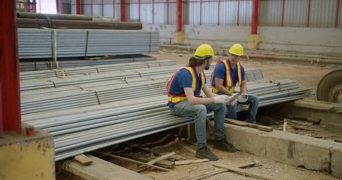 Two industrial workers collaborate while inspecting inventory data on a digital tablet inside a large metal fabrication warehouse during a quality control check.