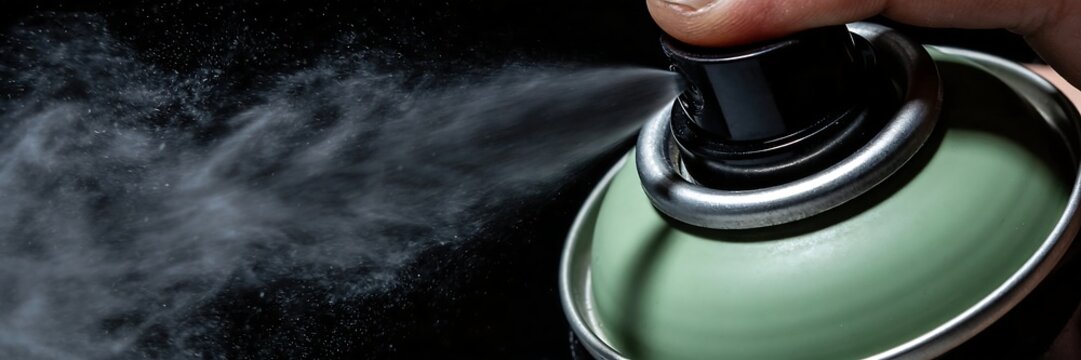 A finger pressed the button on a greenish-gray spray can, spraying out a mist of black liquid.