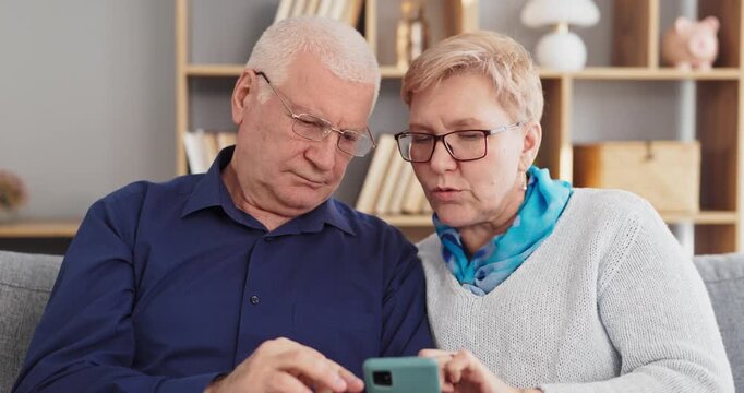 Seniors couple learning smartphone technology together at home. They focus on phone use, getting support for communication and online banking. Concept highlights senior digital help and trust.
