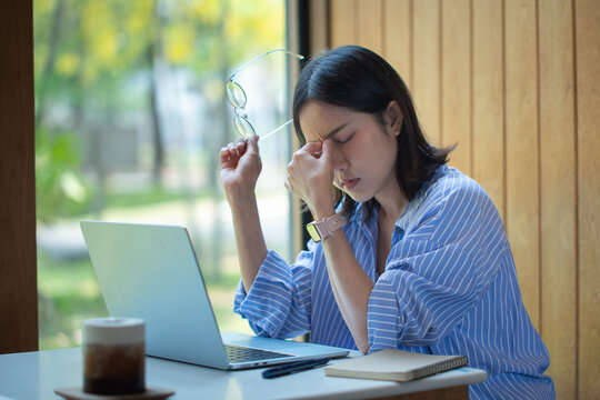 Stressed Asian woman at a cafe rubbing her bridge nose with a headache or eye strain while working on a laptop