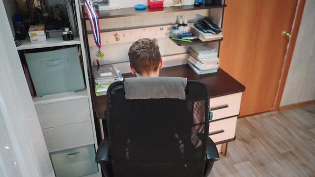 White grandson seated at desk turning gaze sideways, tidy drawers and shelf in background, soft afternoon light, pensive expression suggesting thought and memory, calm household workspace