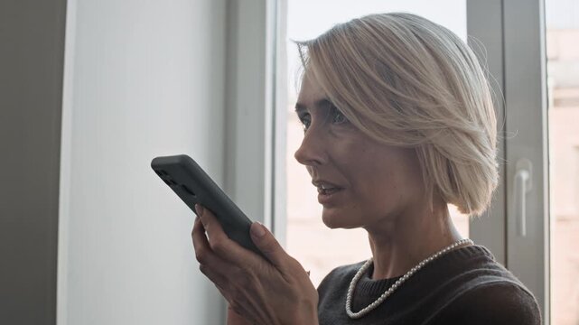 Close up of blonde office worker holding smartphone and recording voice message while working in office