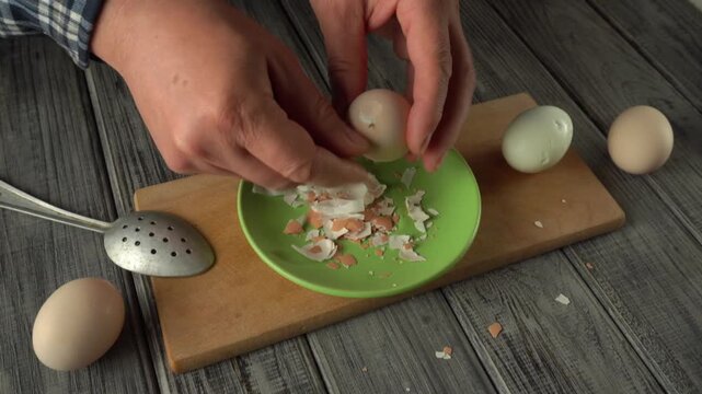 Hands of a man peeling a boiled egg over a green plate, with eggshells and additional eggs arranged on a wooden cutting board in a kitchen setting