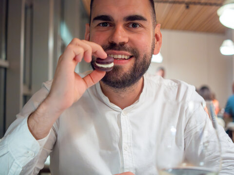 Portrait of a bearded man in a white shirt holding a purple macaron, ready to eat at a restaurant.
