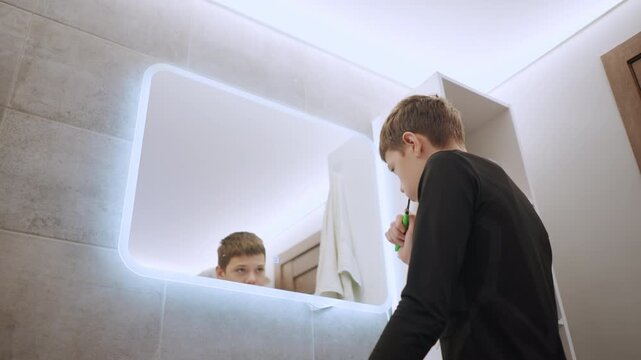 White boy checking reflection at led bathroom mirror while leaning over sink, toothbrush in hand, dim tile surroundings, introspective nighttime mood, modern vanity and toiletries visible
