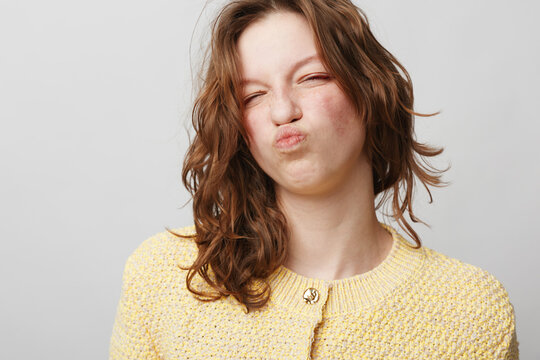 Young woman with curly brown hair wearing yellow sweater makes funny face with puckered lips and closed eyes. Studio portrait on light grey background. Concept of humor and expression.