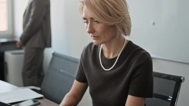 Blonde woman sitting at desk, working on laptop and starting looking at camera, breaking into wide smile. Her colleagues collaborating in background