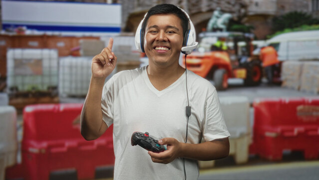 Young man holding game controller points finger upward at construction building outdoors; joy online gaming.