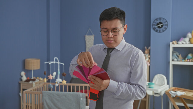 Man latin hispanic wearing shirt and tie holds pink color swatches, pointing with fingers above a wooden cradle in a studio nursery; thoughtful decision.