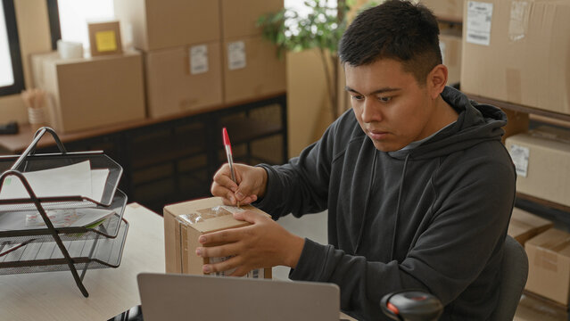 Man writes label on parcel with red pen at laptop desk, hands holding package amid stacked cardboard boxes and organizer trays in building; focused small business shipping.