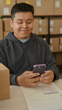 Man holding smartphone with both hands at a packing desk in a building office surrounded by parcels, boxes and paperwork; small business satisfaction.