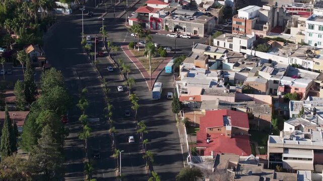 Aerial view of a palm tree lined boulevard with cars and buses in Durango, Mexico. Colorful residential rooftops and urban streets visible with morning shadows and street lights.