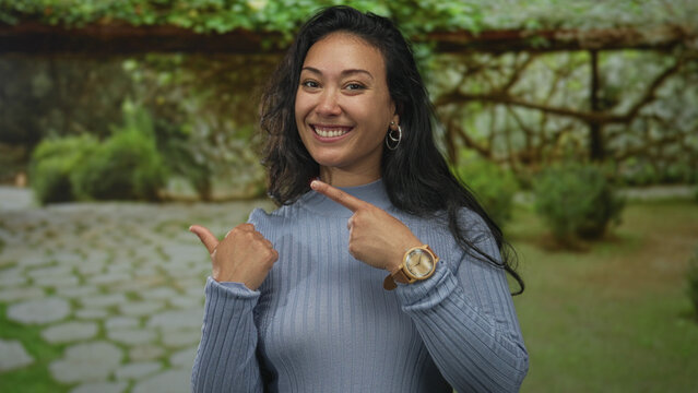 Woman smiling and points thumb to side in forest wearing blue sweater and wooden watch, casual outdoor pose; joy confidence.