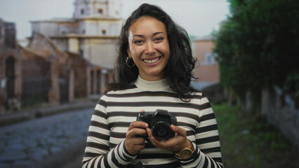 Woman holding camera and smiling, striped sweater and hoop earrings visible, standing on cobblestone street by historic building and trees  joy curiosity. © Krakenimages.com