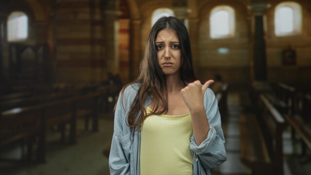 Young woman points thumb over shoulder toward a pew inside church building, frowning with a displeased expression and subtle shoulder lean; annoyance doubt protest.