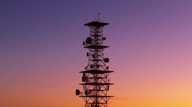 Tall telecommunications tower silhouetted against a stunning gradient sunset sky transitioning from purple to orange. Multiple satellite dishes and antennas with blinking red warning lights visible.