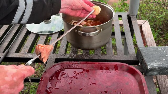 Hands of a cook threading raw marinated meat and onion rings onto a skewer over a preparation station. Metallic pot with marinade and a red tray in the background. Spring picnic concept